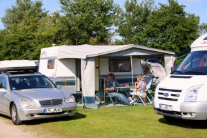 Campingplatz Ausstattung: Stellplatz mit Wohnwagen und Campinggäste, davor ihr PKW nebst Wohnmobil